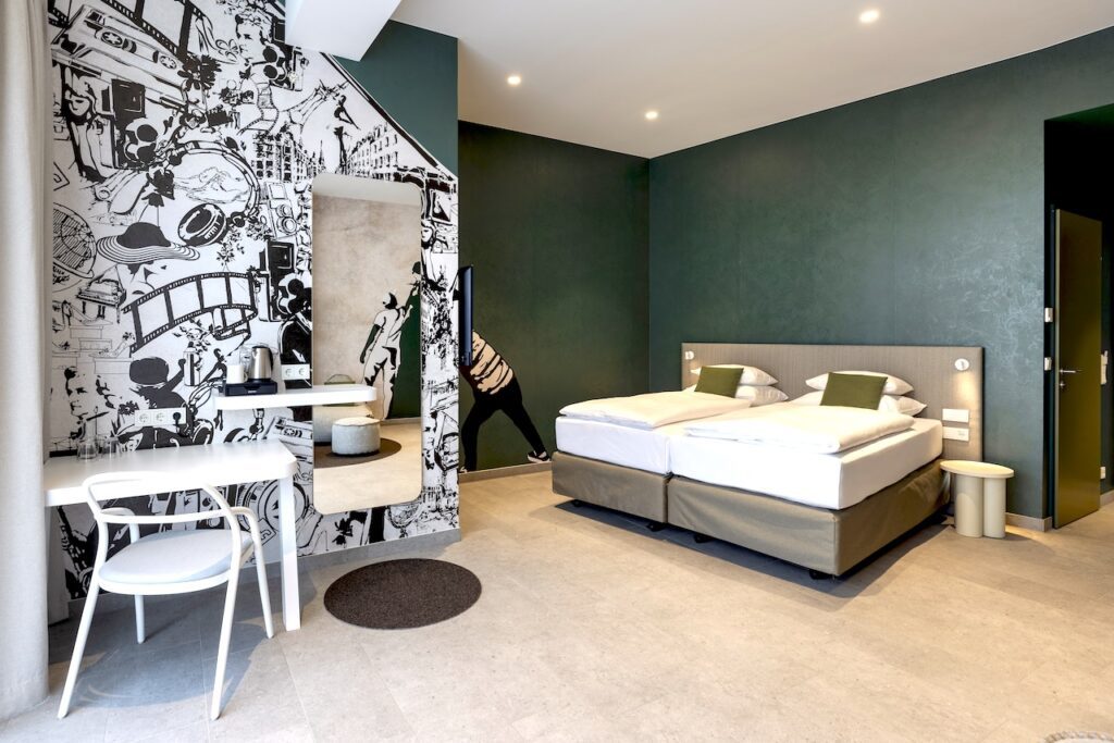 Zimmer im Stzadthotel Arcotel AQ mit Doppelbett, Schreibtisch und einem wandhohen Moderenen Gemälde