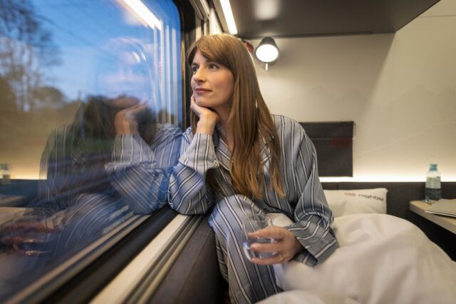 Junge blonde Frau sitzt auf dem Bett in einer Kabine und blickt beim Nachtzug-Fenster in die Landschaft