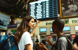 Reisemarkt in Europa: Wie ist denn die Lage? Zwei junge Mädchen am Flughafen. Eines von ihnen zeigt auch eine beleuchtete Anzeigetafel mit den Abflugzeiten