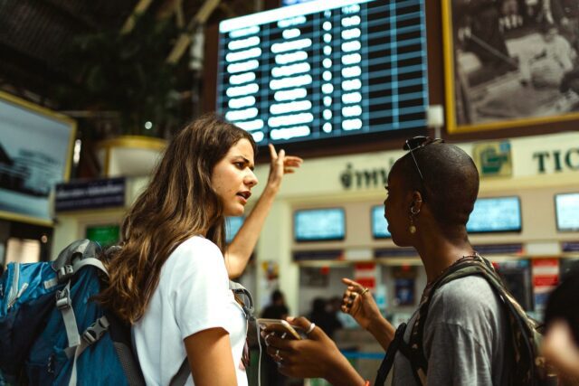 Reisemarkt in Europa Zwei junge Mädchen am Flughafen. Eines von ihnen zeigt auch eine beleuchtete Anzeigetafel mit den Abflugzeiten