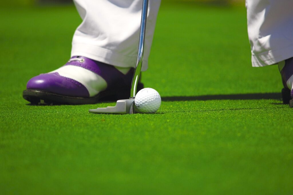 Golfsport begeistert immer mehr Menschen. Grüner Rasen, auf dem ein Golfer mit seinen Golfschuhen steht und mit dem stählernen Butter einen weißen Golfball ins Loch bewegt