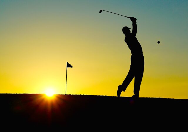 Sonnenuntergang auf einem Golfplatz, ein Mann holt zum Abschlag aus und der Ball als schwarzer Punkt fliegt durch das Licht des Sonnenuntergangs