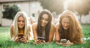 Social Media im Urlaub? Ja, warum nicht! Social Media im Urlaub: Drei junge Frauen liegen auf dem Rasen und durchsuchen ihre Smartphones nach Neuigkeiten aus Social Media
