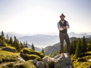 Bayern – direkt, kompetent und persönlich! Ein Mann mit Hut steht auf einem Berg in Bayern mit verschränkten Armen