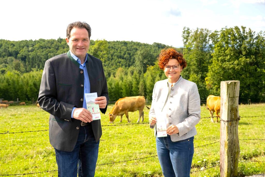 Landwirtschaftsminister Norbert  Totschnig und Tourismus-Staatssekretärin Elisabeth Zehetner stehen vor einem Drahtzaun, im Hintergrund grast Weidevieh. Sie empfehlen 10 Regeln für einen sicheren Umgang mit Weidevieh auf Almen