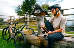Steiermark mit dem Rad und Bike entdecken Steiermark erraten: Junge Frau mit Helm und einer Fasche sitzt auf einem Holztrog, neben ihr zwei Mountainbikes