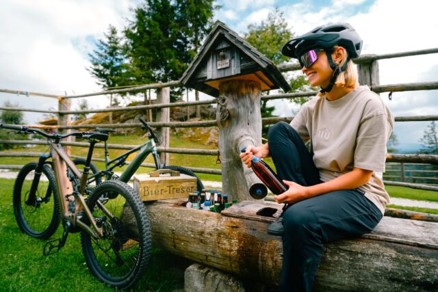 Steiermark entdecken Steiermark erraten: Junge Frau mit Helm und einer Fasche sitzt auf einem Holztrog, neben ihr zwei Mountainbikes