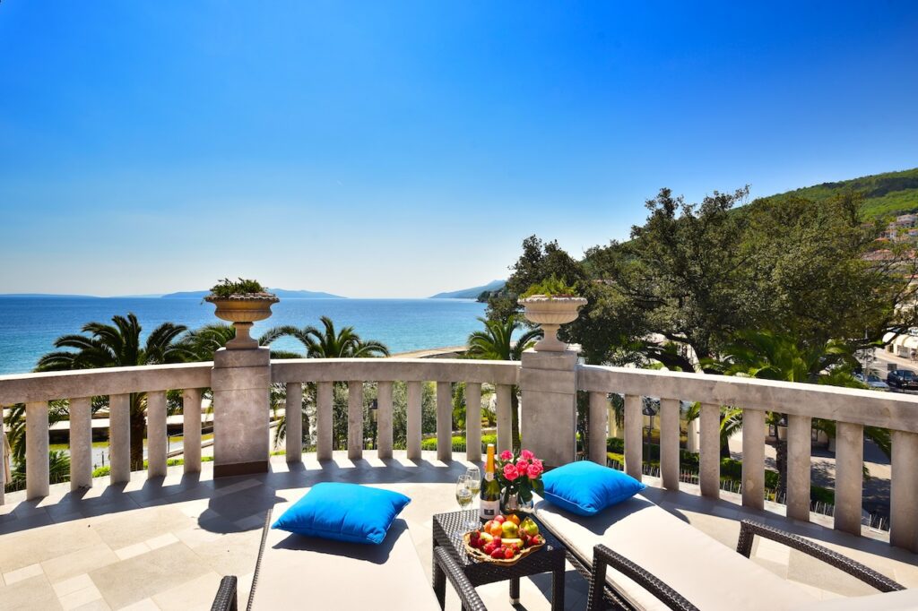 Sonnige Terrasse mit bleiben Sitzpolstern und Blick aufs Meer und bewaldete Berge ringsum