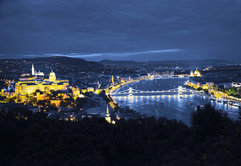 Abendaufnahme von Budapest: Der festlich beleuchtete Burgpalast, in der Mitte die Kettenbrücke und dahinter das Parlament im gotischen Stil