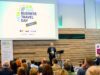 Business Travel Day Connect in Wien Connect Day: Ein Mann steht an einem Pult vor vielen Menschen und hält eine Rede. Im Hintergrund das Logo von ABTA eingeblendet