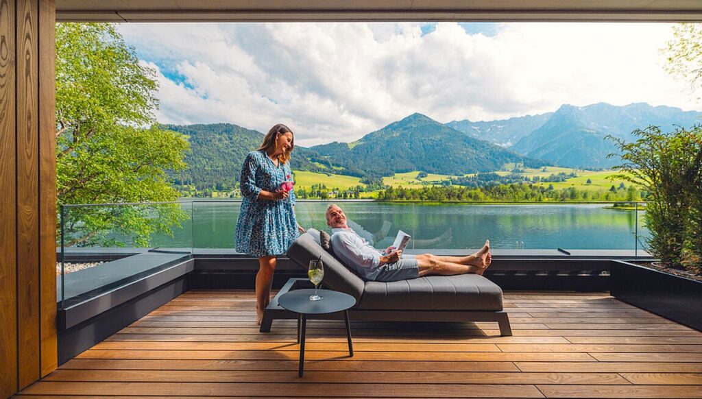Hotels für Firmenevents: Ein Mann auf einer Liege, hinter ihm steht eine Frau, großes Fenster mit Blick auf einen See