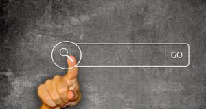 Wie digitale Quellen Urlauber beeinflussen Digitale Quellen als Helfer: Vor einer dunklen Schiefertafel zeigt der Daumen einer Hand auf ein Suchfeld