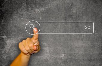 Wie digitale Quellen Urlauber beeinflussen Digitale Quellen als Helfer: Vor einer dunklen Schiefertafel zeigt der Daumen einer Hand auf ein Suchfeld