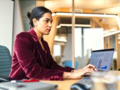 So reisen Geschäftsreisende sparsam mit KI Das Reisen für Geschäftsreisende verändert sich laufend, seit Digitalisierung und KI im Travel Management eingesetzt werden: Eine Frau in roter Bluse sitzt vor einem Laptop im Büro