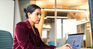 So reisen Geschäftsreisende sparsam mit KI Das Reisen für Geschäftsreisende verändert sich laufend, seit Digitalisierung und KI im Travel Management eingesetzt werden: Eine Frau in roter Bluse sitzt vor einem Laptop im Büro