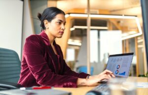 So reisen Geschäftsreisende sparsam mit KI Das Reisen für Geschäftsreisende verändert sich laufend, seit Digitalisierung und KI im Travel Management eingesetzt werden: Eine Frau in roter Bluse sitzt vor einem Laptop im Büro