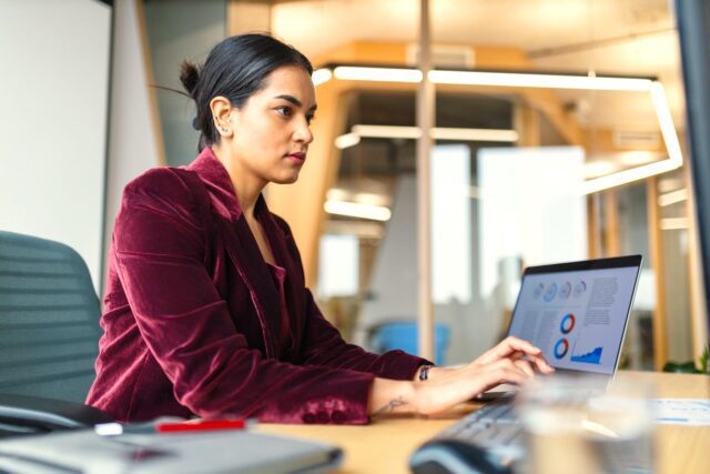 Reisen in Zukunft Das Reisen für Geschäftsreisende verändert sich laufend, seit Digitalisierung und KI im Travel Management eingesetzt werden: Eine Frau in roter Bluse sitzt vor einem Laptop im Büro