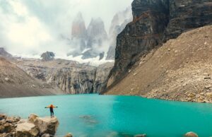 Chile: Ziel für Abenteurer und Naturfreunde Zwischen Bergen, Gletschern und endlosem Himmel erstrecken sich in Chile die Torres del Paine, die „Türme des blauen Himmels“, im Nationalpark : Eine Person steht auf einem Felsvorsprung und blickt auf den grünen See mit dem nebelverhangenen Schneegipferln