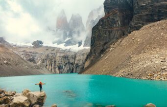 Chile: Ziel für Abenteurer und Naturfreunde Zwischen Bergen, Gletschern und endlosem Himmel erstrecken sich in Chile die Torres del Paine, die „Türme des blauen Himmels“, im Nationalpark : Eine Person steht auf einem Felsvorsprung und blickt auf den grünen See mit dem nebelverhangenen Schneegipferln