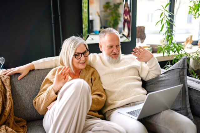 Ein älteres Paar – Best Ager – sitzt lässig auf einem Sofa und blickt auf das Display eines Laptops