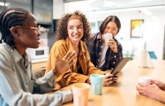 So ticken Gen Z und Boomer auf Geschäftsreisen Gen Z und Boomer: Junge Frauen und ein dunkelhäutiger Mann sitzen an einem Tisch und unterhalten sich