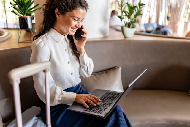 Routinen machen Geschäftsalltag flexibel: Frau mit Trollen sitzt in einem Büro, hält ein Mobiltelefon ans Ohr und tippt auf dem Keyboard ihres Laptops
