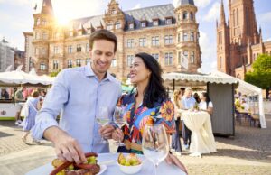 Wiesbaden 2026: Kultur, Wein & Hollegha Ein Mann und ein Frau in ei9nem bunten Kleid Besuchern die Rheingauer Weinwoche in Wiesbaden