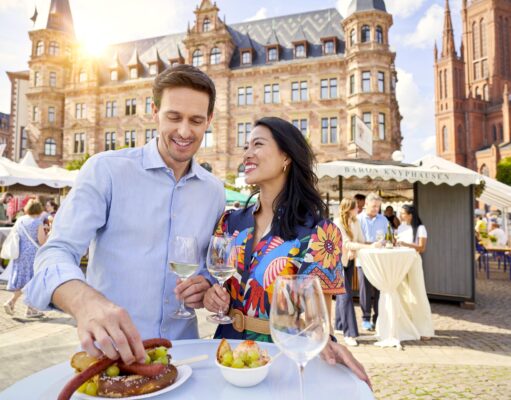Wiesbaden 2026: Kultur, Wein & Hollegha Ein Mann und ein Frau in ei9nem bunten Kleid Besuchern die Rheingauer Weinwoche in Wiesbaden