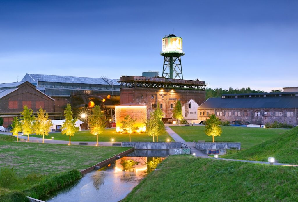 Die Jahrhunderthalle in Bochum in den Abendstunden. Davor befinden sich Industriegebäude und Rasenflächen. Westpark in Bochum: Industrie als Objekt auf der IGA 2027 Ruhrgebiet wird (Bild: Francesco Carovillano, DZT)