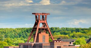 DZT vermarktet IGA 2027 Ruhrgebiet global Eine große, rote Stahlkonstruktion namens Doppelbock Förderturm der Zeche Zollverein in Essen, die Teil eines Industriekomplexes mit Backsteingebäuden ist. Dichte grüne Bäume füllen den Hintergrund unter einem bewölkten Himmel.