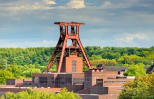 DZT vermarktet IGA 2027 Ruhrgebiet global Eine große, rote Stahlkonstruktion namens Doppelbock Förderturm der Zeche Zollverein in Essen, die Teil eines Industriekomplexes mit Backsteingebäuden ist. Dichte grüne Bäume füllen den Hintergrund unter einem bewölkten Himmel.