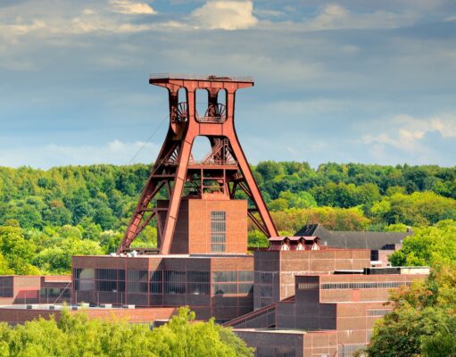 DZT vermarktet IGA 2027 Ruhrgebiet global Eine große, rote Stahlkonstruktion namens Doppelbock Förderturm der Zeche Zollverein in Essen, die Teil eines Industriekomplexes mit Backsteingebäuden ist. Dichte grüne Bäume füllen den Hintergrund unter einem bewölkten Himmel.