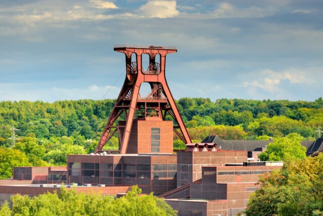 Eine große, rote Stahlkonstruktion namens Doppelbock Förderturm der Zeche Zollverein in Essen, die Teil eines Industriekomplexes mit Backsteingebäuden ist. Dichte grüne Bäume füllen den Hintergrund unter einem bewölkten Himmel.