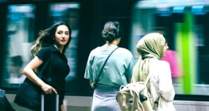 Travel Risk Awareness: 7 Risiken für Geschäftsreisen Travel Risk Awareness auf Geschäftsreisen: Junge Frau mit schwarzem Koffer, Bahnhof, vorbeifahrender Zug, zwei Reisende neben ihr