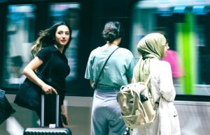 Travel Risk Awareness: 7 Risiken für Geschäftsreisen Travel Risk Awareness auf Geschäftsreisen: Junge Frau mit schwarzem Koffer, Bahnhof, vorbeifahrender Zug, zwei Reisende neben ihr