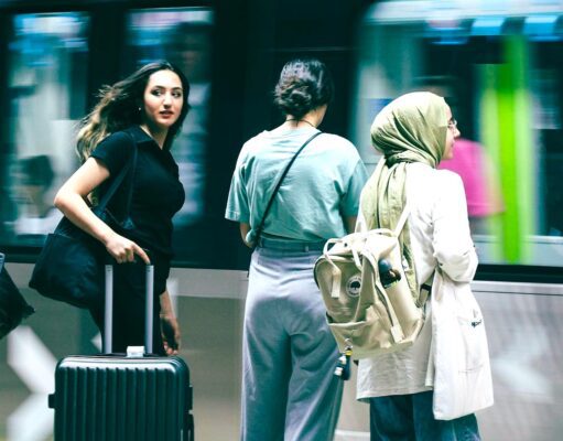 Travel Risk Awareness: 7 Risiken für Geschäftsreisen Travel Risk Awareness auf Geschäftsreisen: Junge Frau mit schwarzem Koffer, Bahnhof, vorbeifahrender Zug, zwei Reisende neben ihr