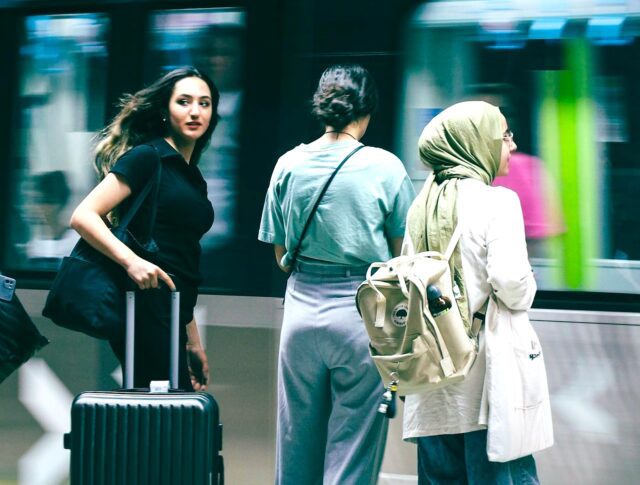 Travel Risk Awareness auf Geschäftsreisen: Junge Frau mit schwarzem Koffer, Bahnhof, vorbeifahrender Zug, zwei Reisende neben ihr