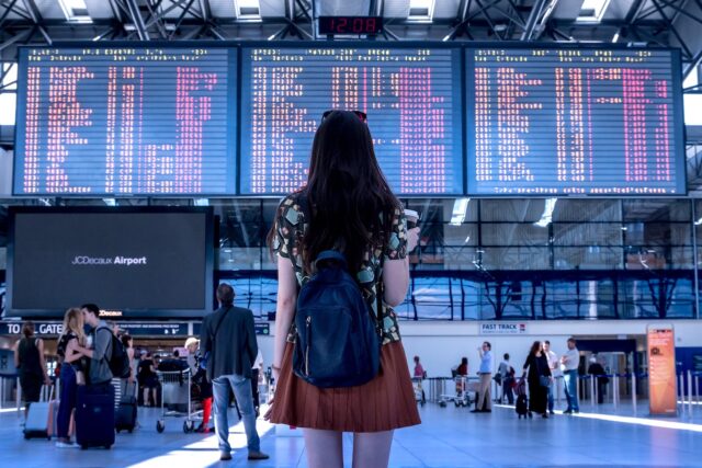 Social Media und Reiseentscheidungen: junge frau mit rucksack blickt auf eine große anzeigetafel mit abflügen in Flughafen