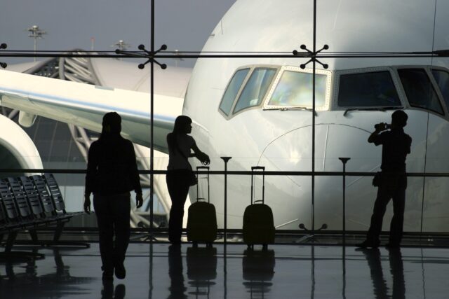 Wie wichtig sind Geschäftsreisen? Wie wichtig sind Geschäftsreisen wirklich? Eine Frau mit Handgepäck blickt von der Abflughalle durch einen großes Fenster auf ein Flugzeug