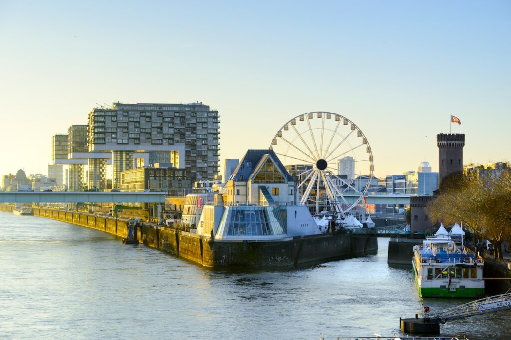 Incoming Tourismus in Deutschland: Rheinauhafen mit Kranhäusern, Riesenrad und Weihnachtsmarktständen entlang der Promenade am Rhein bei warmem Licht und Blick auf Wasser und Uferarchitektur
