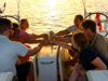 Kojencharter: Segelurlaub für neue Zielgruppen Kojencharter: Segelurlaub für neue Zielgruppen: Vier Personen sitzen in einem Segelboot, im Higergrund spiegelt sich die Sonne im Meer