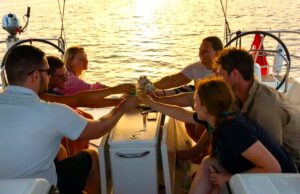 Kojencharter: Segelurlaub für neue Zielgruppen Kojencharter: Segelurlaub für neue Zielgruppen: Vier Personen sitzen in einem Segelboot, im Higergrund spiegelt sich die Sonne im Meer