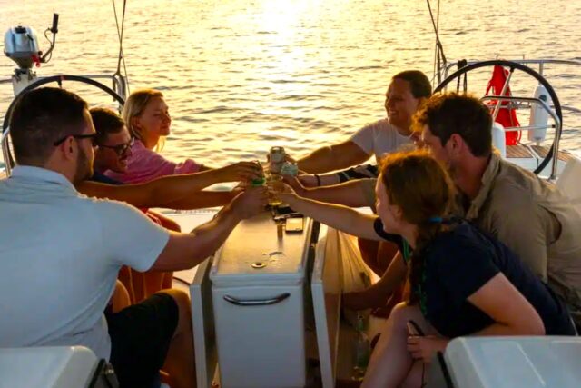 Kojencharter: Segelurlaub für neue Zielgruppen Kojencharter: Segelurlaub für neue Zielgruppen: Vier Personen sitzen in einem Segelboot, im Higergrund spiegelt sich die Sonne im Meer