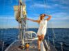 Segeln im Wandel: Mehr Frauen als Skipper Segeln im Wandel: Mehr Frauen als Skipper. Eine Frau im weißen Kleid und mit Sonnenbrille hält sich am, Mast eines Segelschiffes fest und blickt in die Sonne