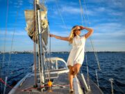Segeln im Wandel: Mehr Frauen als Skipper Segeln im Wandel: Mehr Frauen als Skipper. Eine Frau im weißen Kleid und mit Sonnenbrille hält sich am, Mast eines Segelschiffes fest und blickt in die Sonne