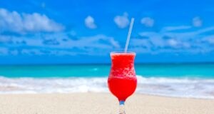Karibik-Regionalnetz der Air France erweitert Karibik-Regionalnetz der Air France erweitert: Willkommen in Übersee. Auf weißem Sandstrand, im Hintergrund das blaue Meer und der blauen Himmel, steht ein Glas mit pinken Getränk und Strohhalm