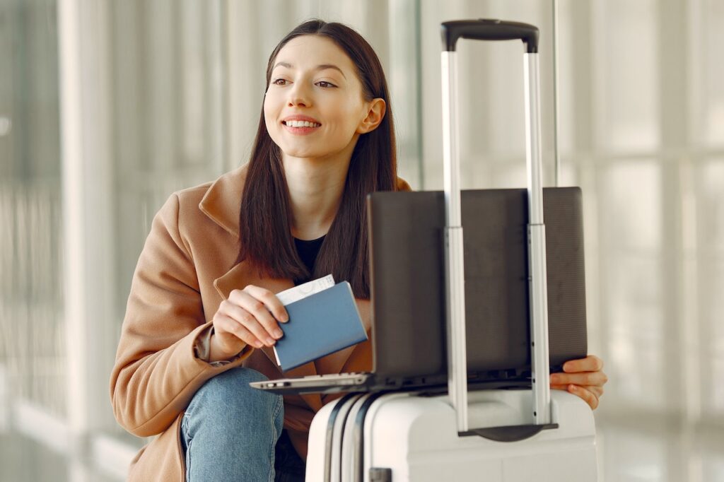 Neue Rolle der Travel Manager: Eine Frau mit Pass in der Hand und einem Metall-Trolley wartet sitzend auf Abflug.