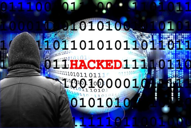 Cyberangriffe auf Reisebüros Cyberangriff auf Reisebüros: Ein Mann im Mantel und ein Monitor mit dem roten Titel „Hacker“ sowie verschiedene Daten im Hintergrund symbolisieren einen Cyberangrifdfd von einem Hacker
