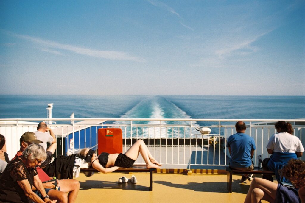 Passagiere auf dem sonnigen Deck eines Schaffes, auf einer Bank liegt eine junge Frau, links und rechts Passagiere: Slow Travel ist in