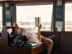 Wie Slow Travel das Urlaubsverhalten prägt Slow Travel: Ein Mann liegt auf einer Sitzbank im Zug, hat ein Buch in der Hand und blickt durch das große Fenster in die Landschaft